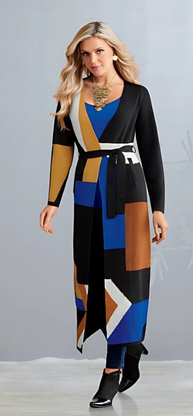 A woman in a colorful, patterned long cardigan and blue top, accessorized with a statement necklace and stylish boots.