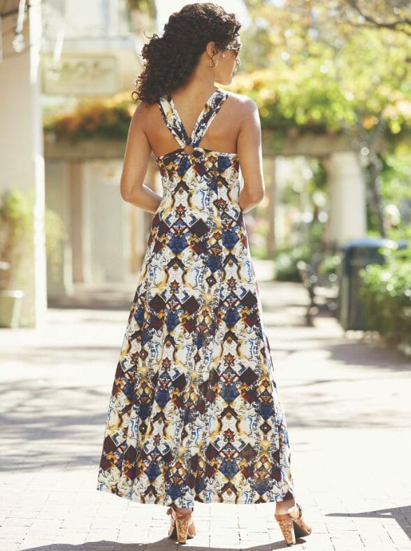 A back view of a woman with long curly hair, wearing a halter style long dress in a white, navy, brown and tan print.