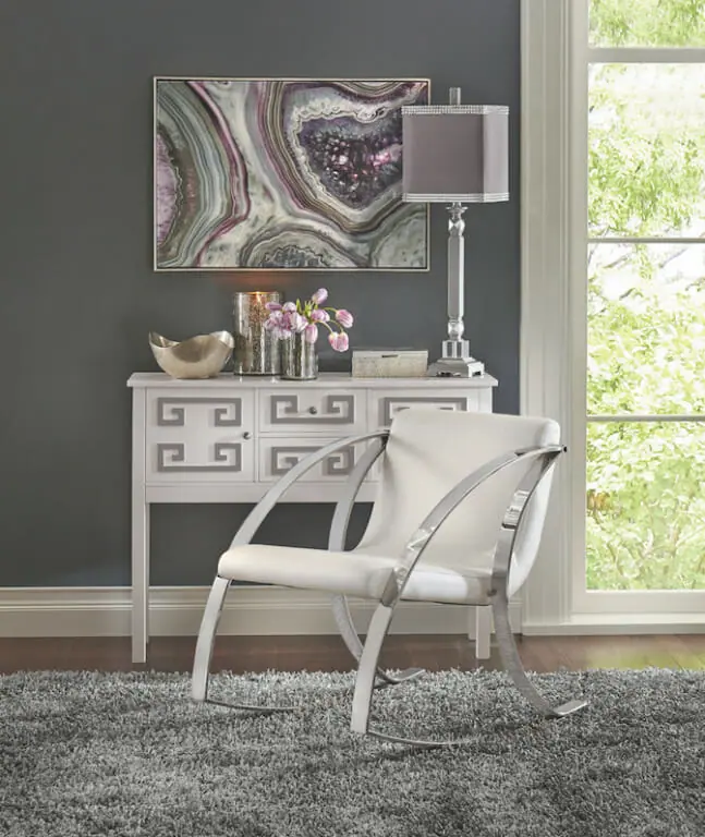 A wall canvas of a purple geode above a white and silver console, a chrome and white chair, and a silver table lamp.