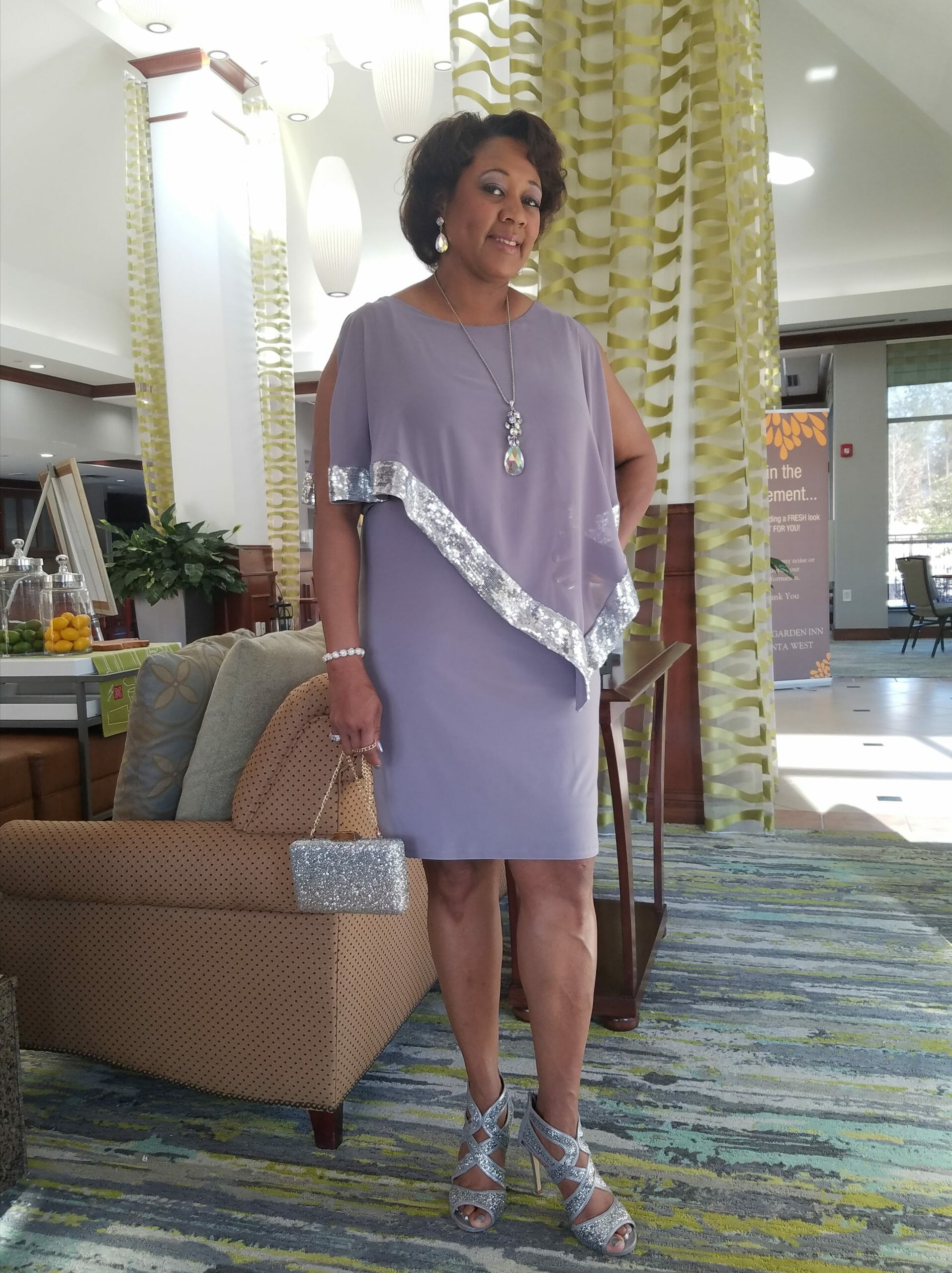 A smiling Midnight Velvet customer, in a lavender dress with silver trim on the cape-like top, and silver heeled sandals.