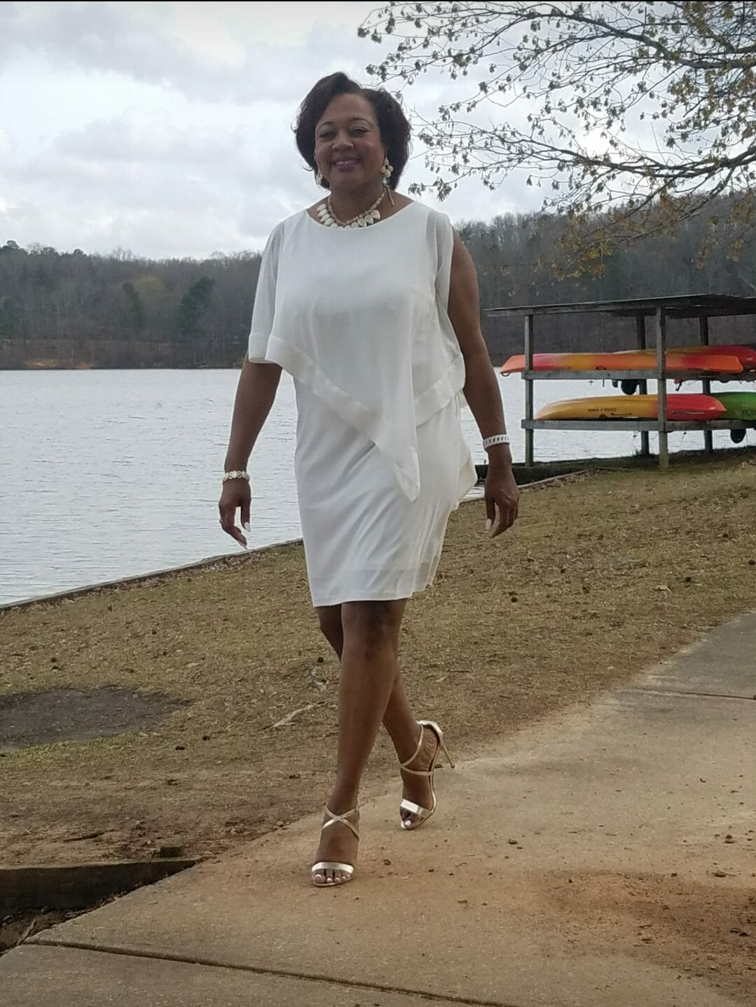 A smiling Midnight Velvet customer outside, in a white dress with satin trim on the cape-like top, and silver sandals.