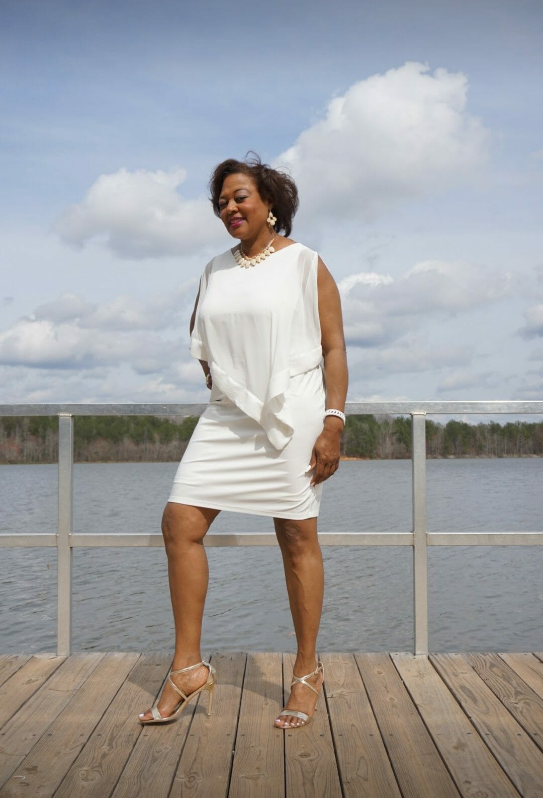 A smiling Midnight Velvet customer lakeside, in a white dress with satin trim on the cape-like top, and silver sandals.