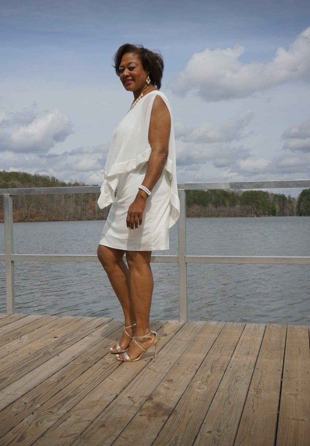 Side view of a smiling Midnight Velvet customer, in a white dress with satin trim on the cape-like top, and silver sandals.