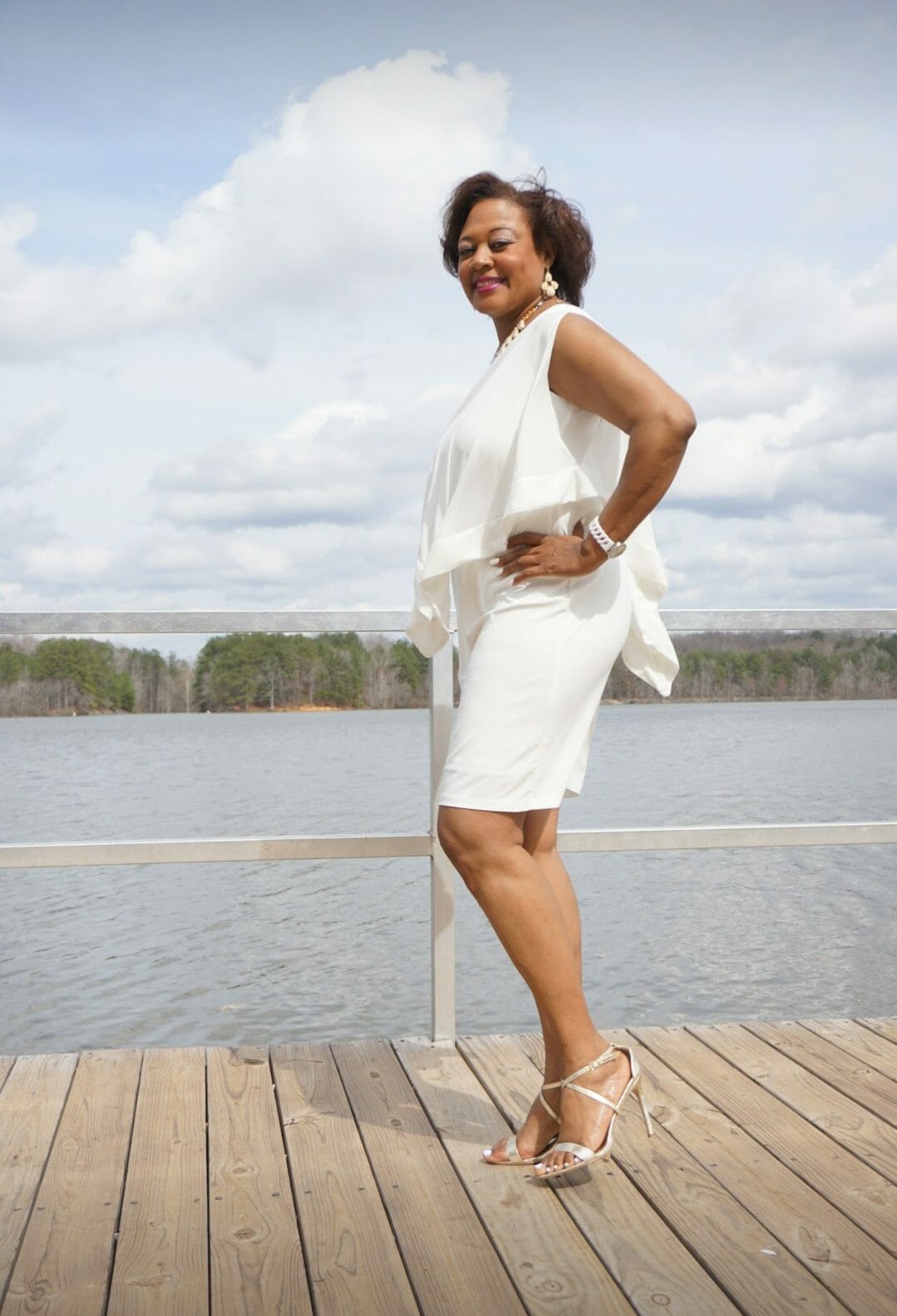 Side view of a smiling Midnight Velvet customer, in a white dress with satin trim on the cape-like top, and silver sandals.