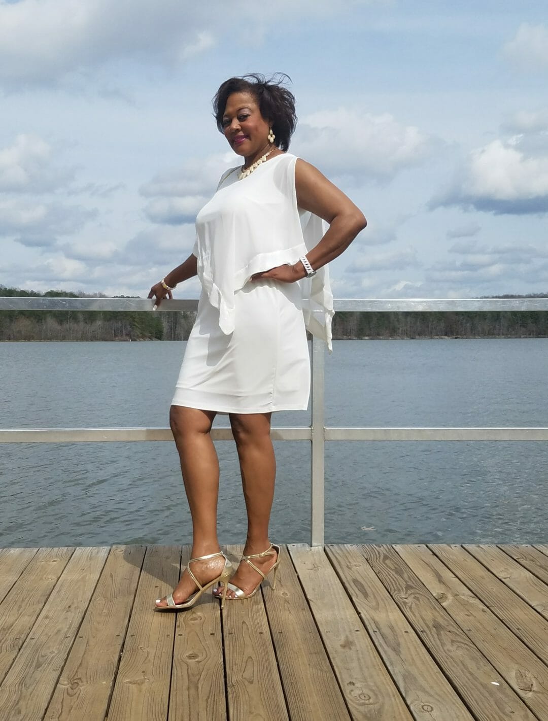 Side view of a smiling Midnight Velvet customer, in a white dress with satin trim on the cape-like top, and silver sandals.