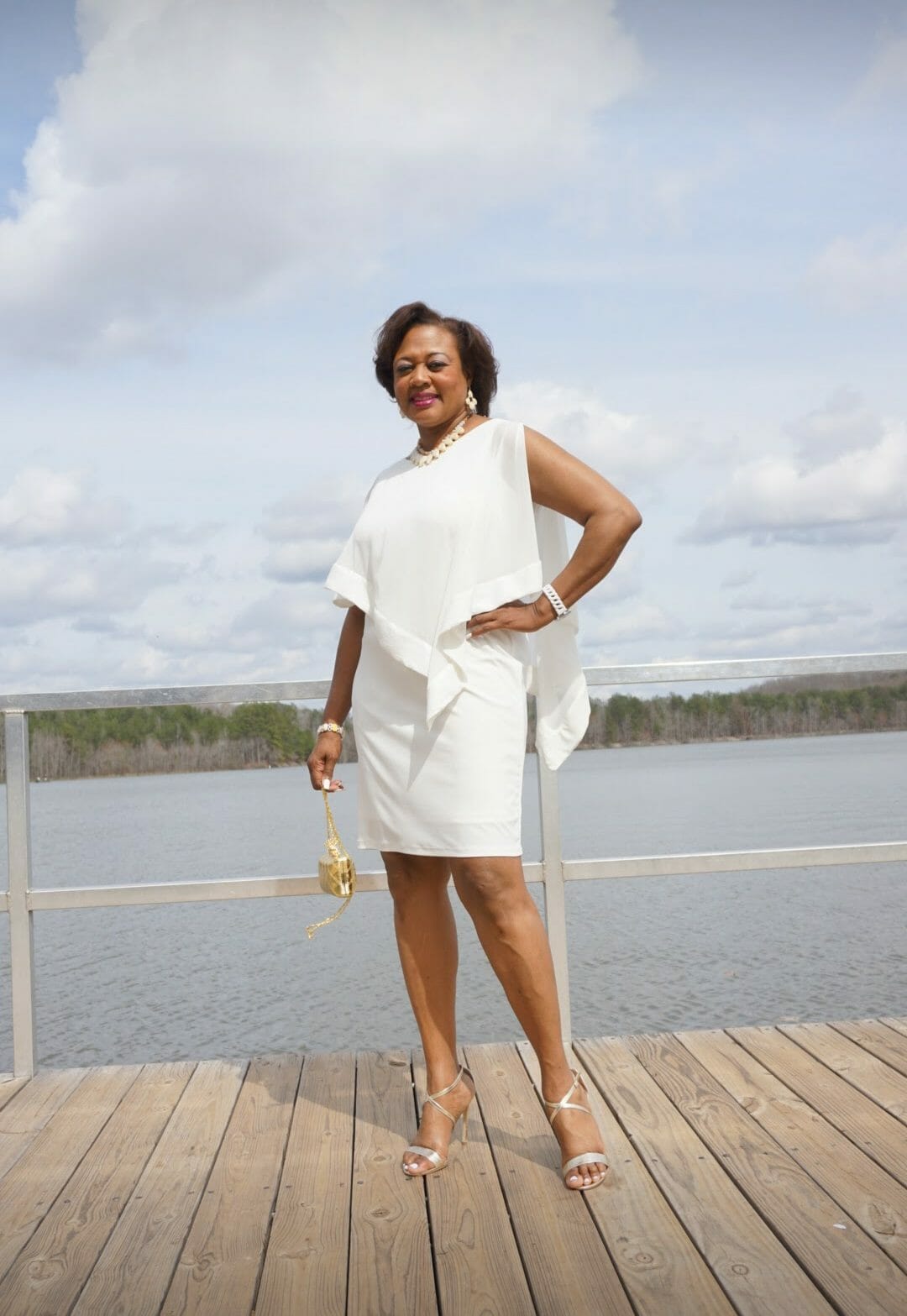 A smiling Midnight Velvet customer lakeside, in a white dress with satin trim on the cape-like top, and silver sandals.