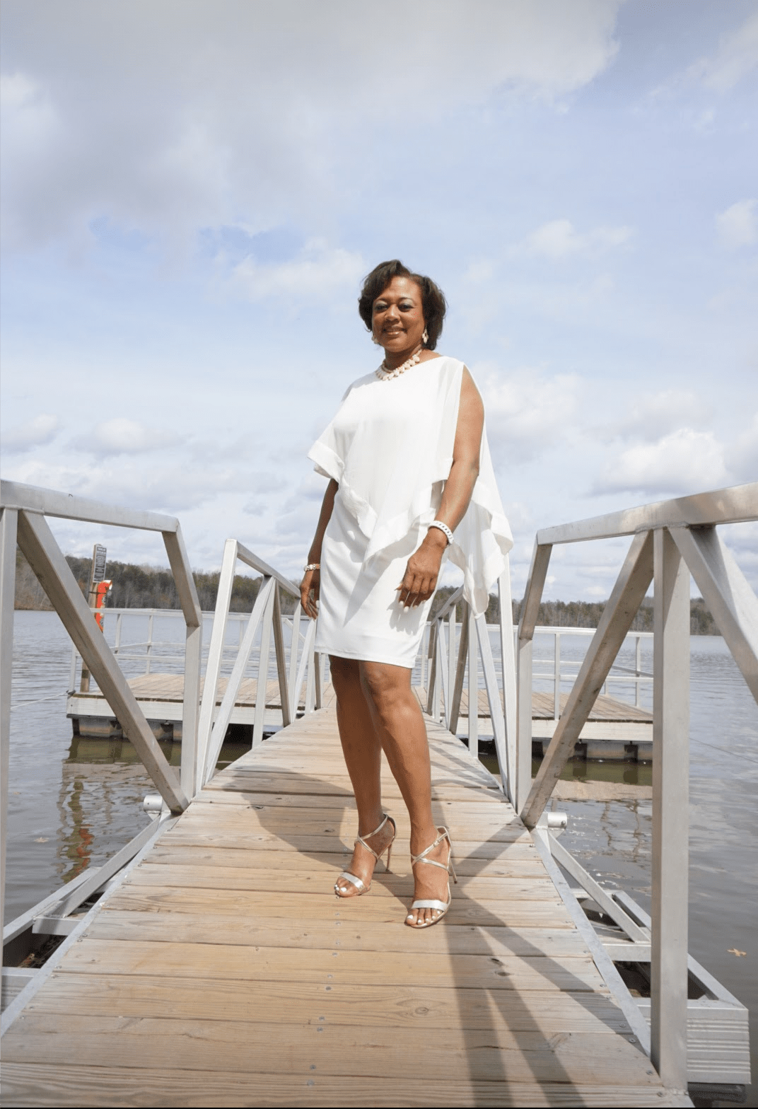 A smiling Midnight Velvet customer on a pier, in a white dress with satin trim on the cape-like top, and silver sandals.