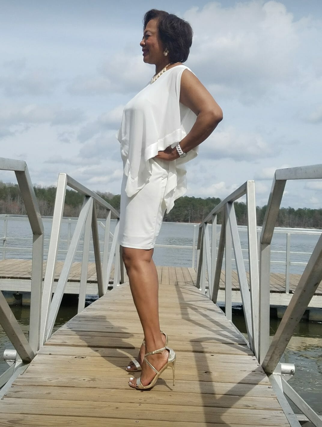 Side view of a smiling Midnight Velvet customer, in a white dress with satin trim on the cape-like top, and silver sandals.