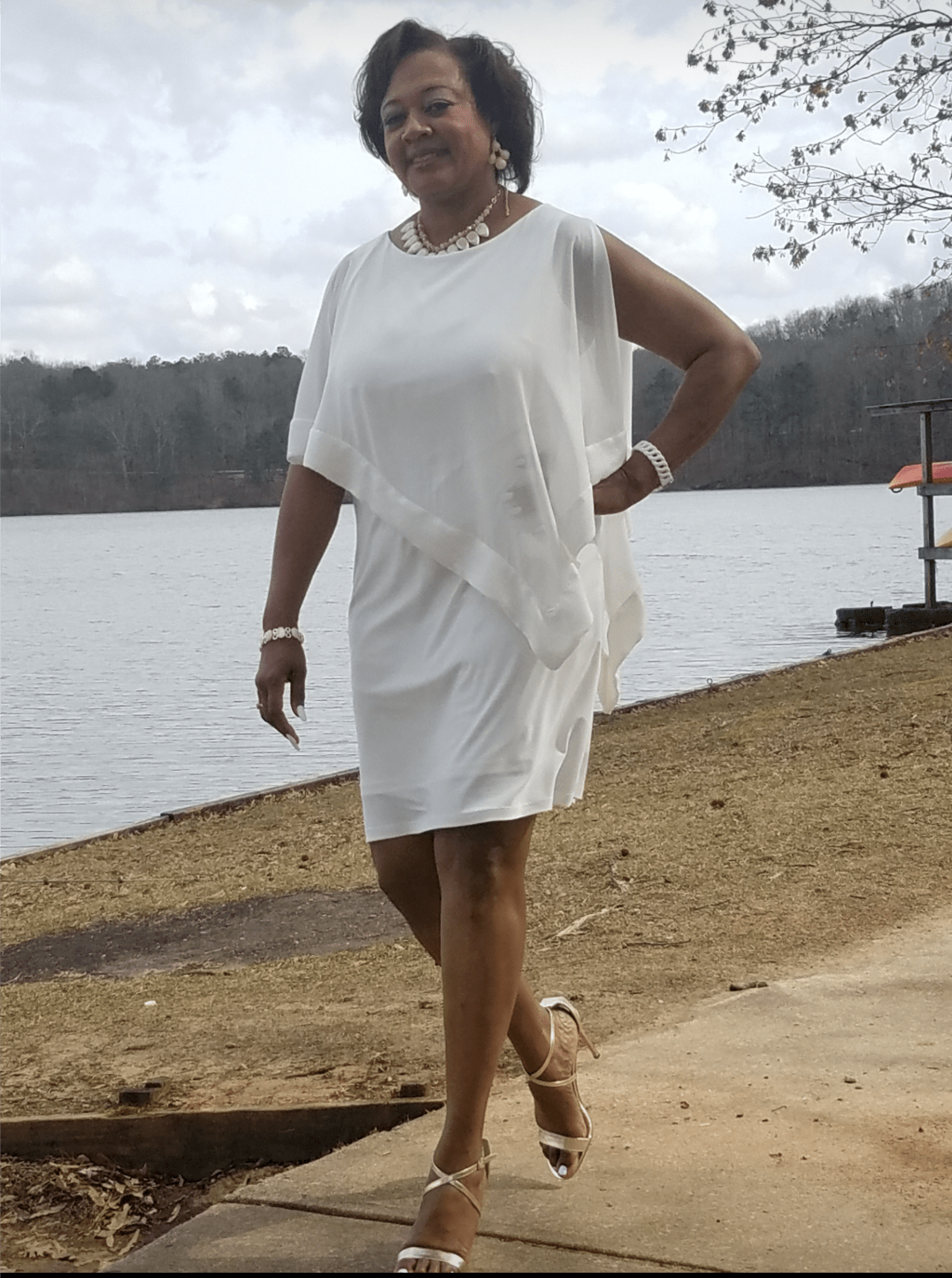 A smiling Midnight Velvet customer lakeside, in a white dress with satin trim on the cape-like top, and silver sandals.