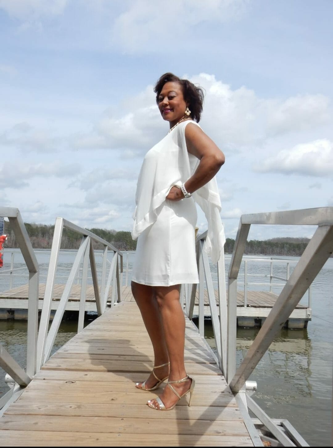 Side view of a Midnight Velvet customer on a pier, in a white dress with satin trim on the cape-like top, and silver sandals.