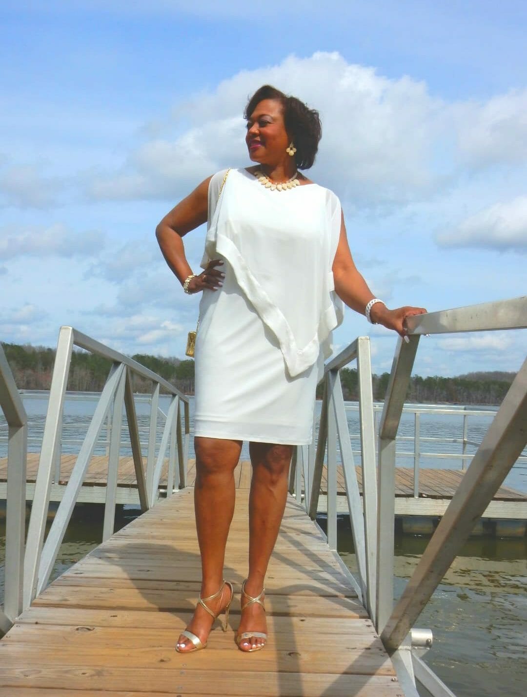 A Midnight Velvet customer on a pier, in a white dress with satin trim on the cape-like top, and silver heeled sandals.