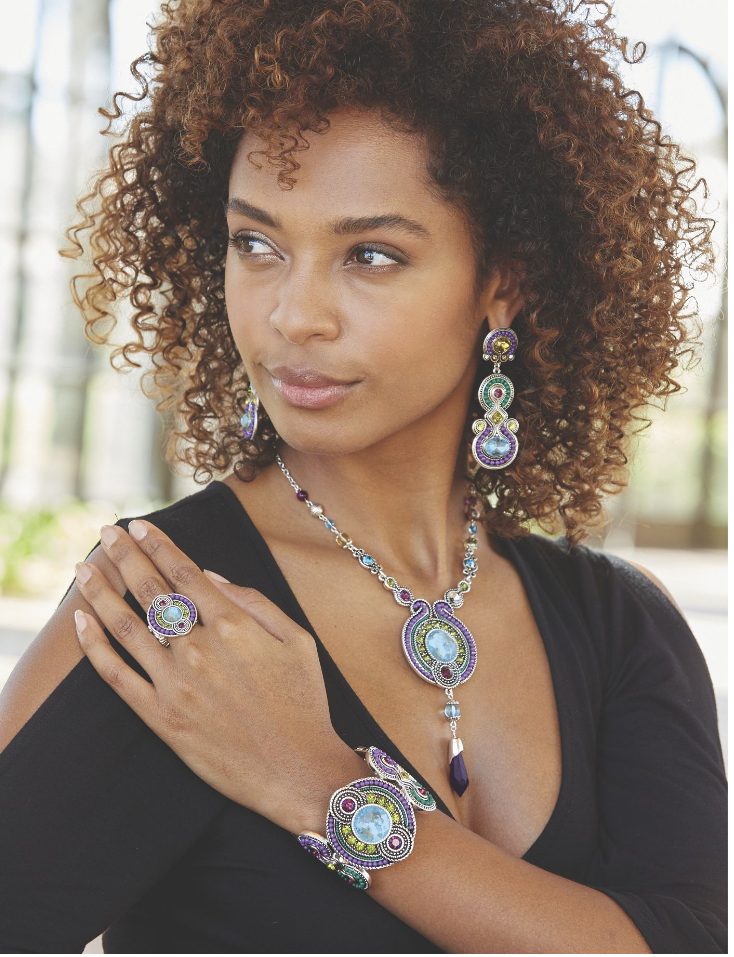 A black woman with curly hair wearing a multi-color Afrocentric design necklace, bracelet, ring, and drop earrings.