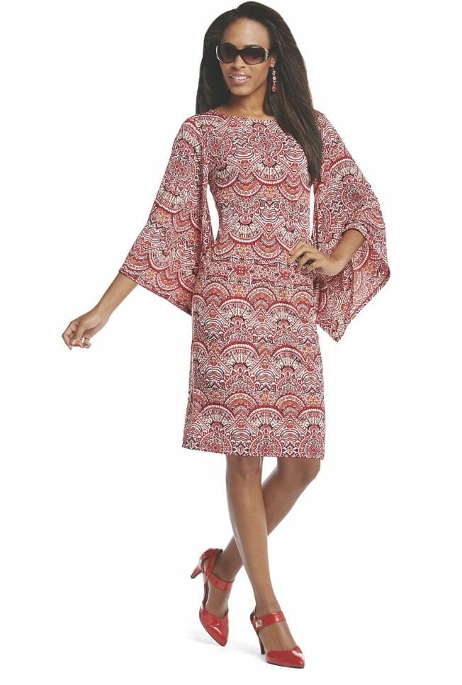 A long-haired black woman in sunglasses and red shoes, wearing a red and white baroque print dress with wide sleeves.