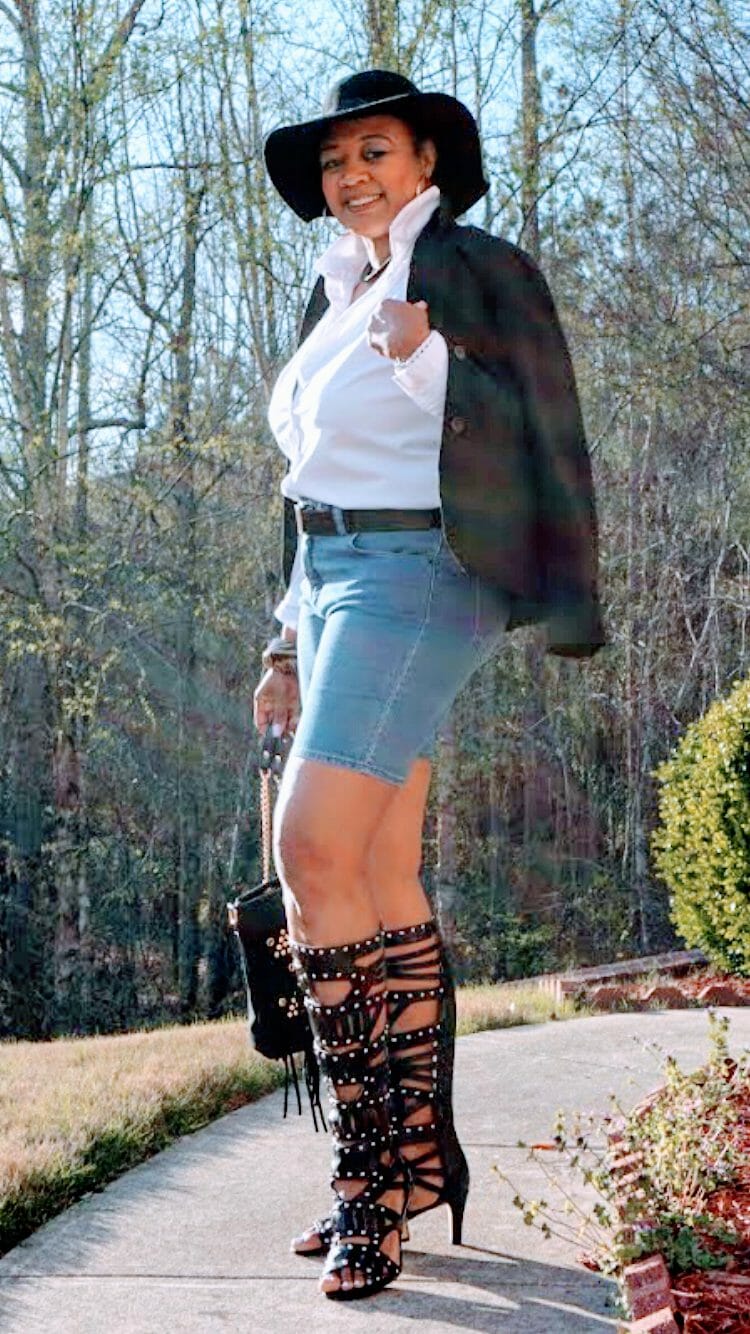 A smiling Midnight Velvet customer in a white shirt, dark jacket, jean shorts, black studded boots, and a brimmed black hat.