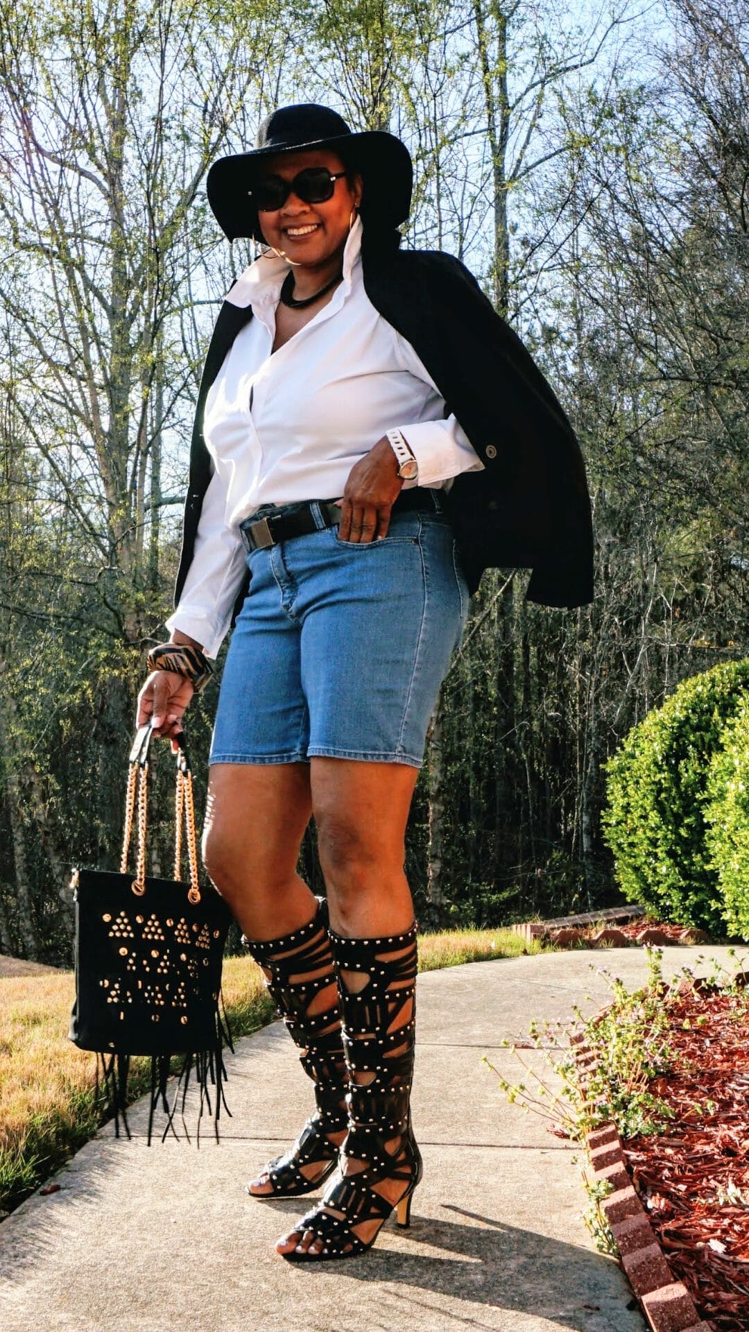 A Midnight Velvet customer in a white shirt and black jacket, jean shorts, black studded boots and bag, and a black hat.
