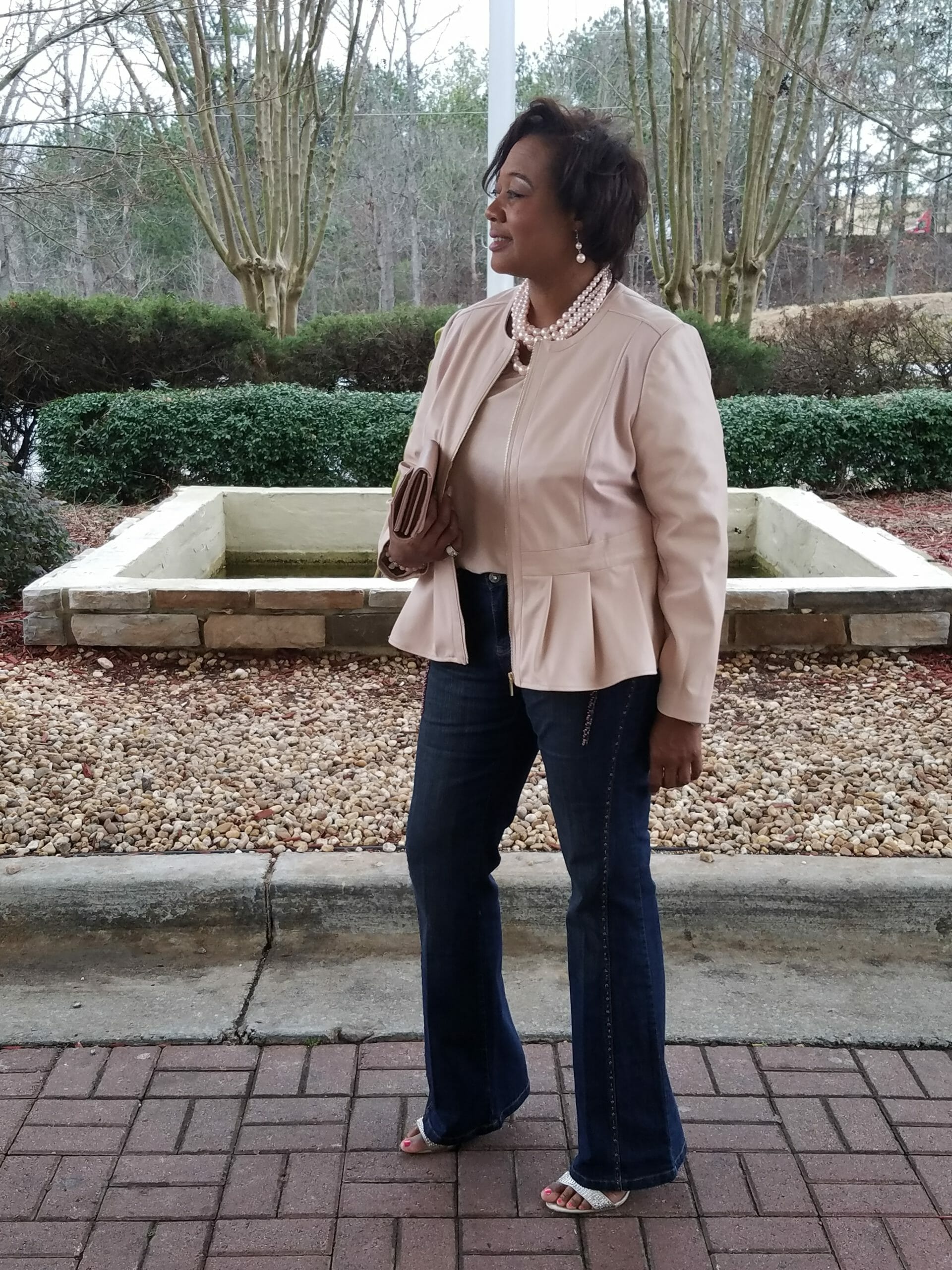 A Midnight Velvet customer outside in a blush top and peplum jacket set, jeans, a pearl necklace, with a blush clutch.