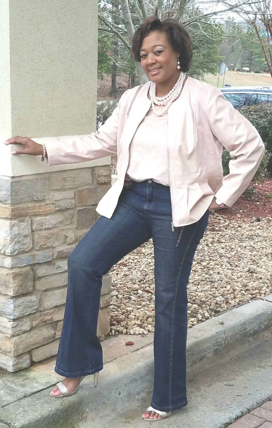 A Midnight Velvet customer by a rock column, in a blush top and peplum jacket set, jeans, sandals, and a pearl necklace.
