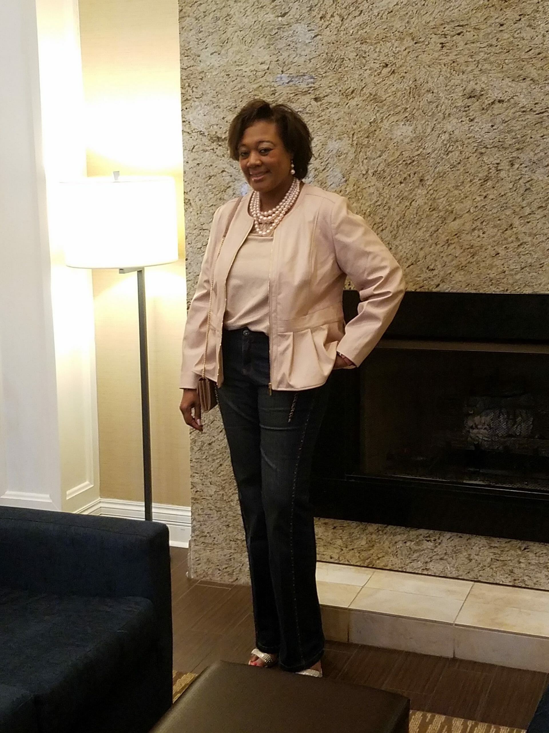 A Midnight Velvet customer by a fireplace, in a blush top and peplum jacket set, jeans, and a pearl necklace.