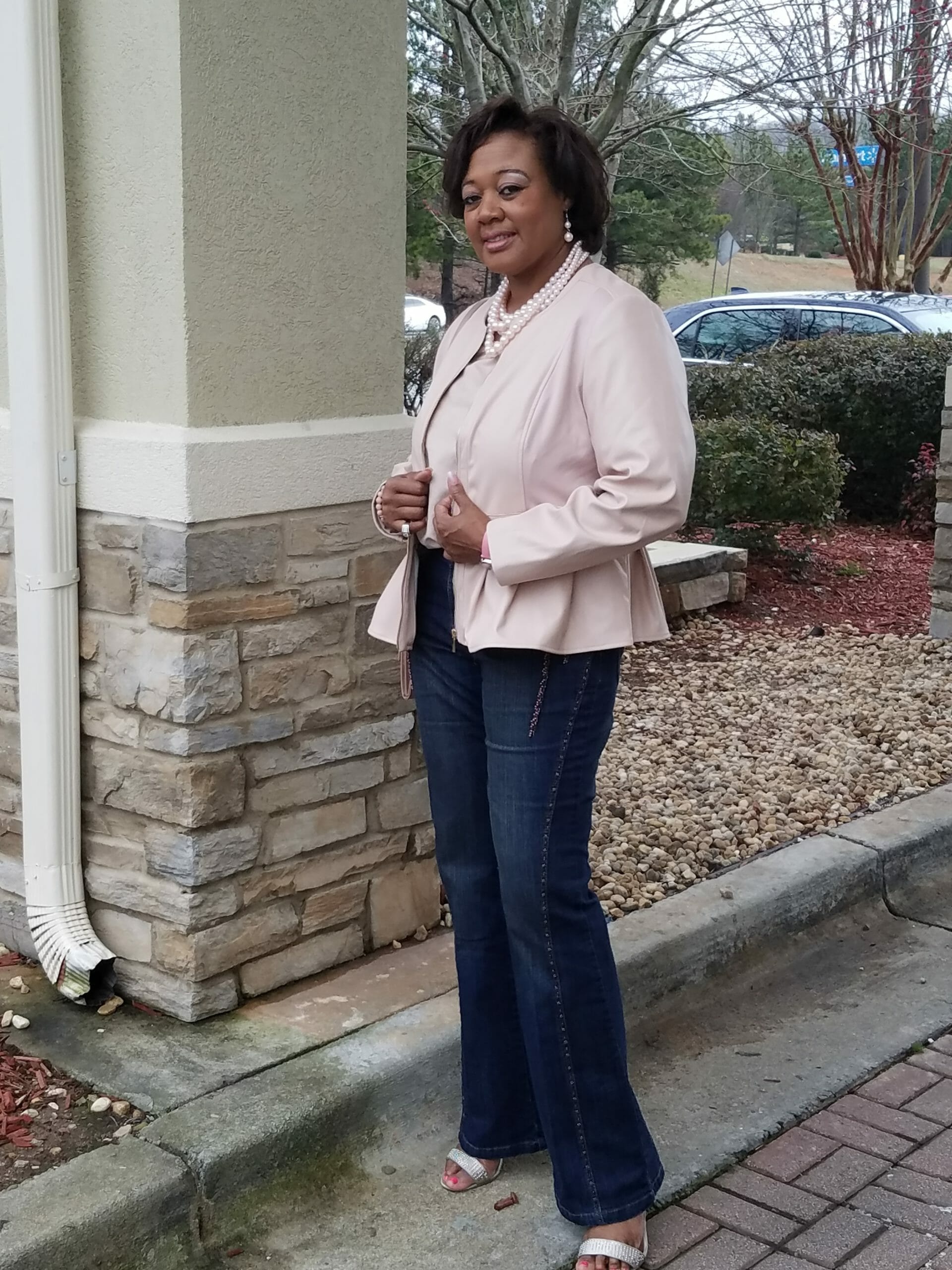 A Midnight Velvet customer by a rock column, in a blush top and peplum jacket set, jeans, sandals, and a pearl necklace.