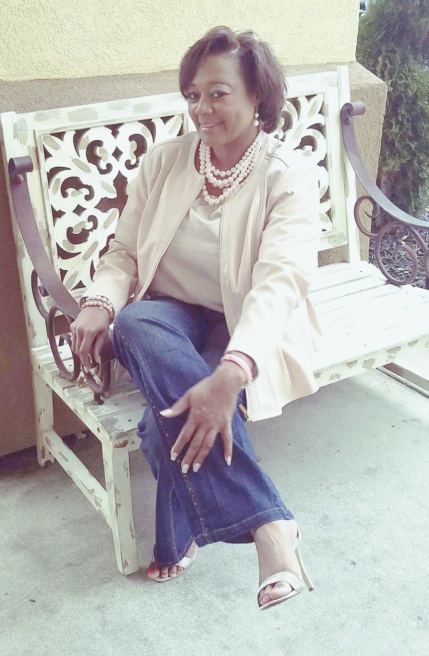 A Midnight Velvet customer by a rock column, in a blush top and peplum jacket set, jeans, sandals, and a pearl necklace.