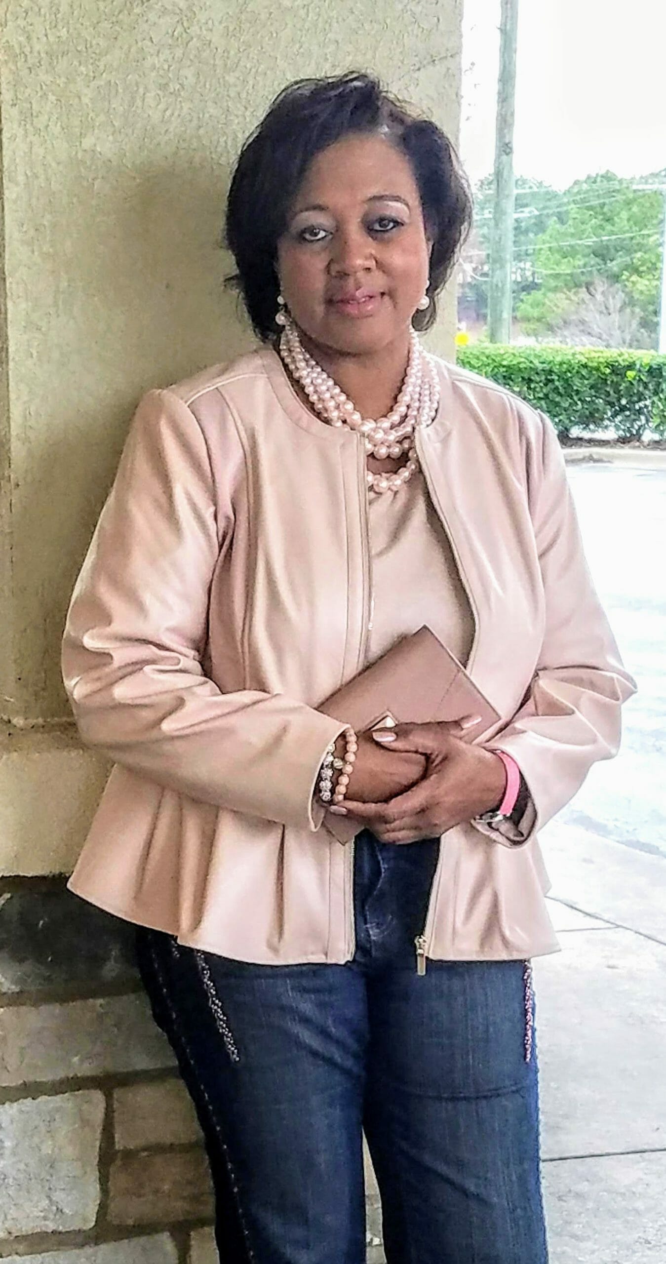 A Midnight Velvet customer in a blush top and peplum jacket set, jeans, a pearl necklace, and holding a blush clutch.