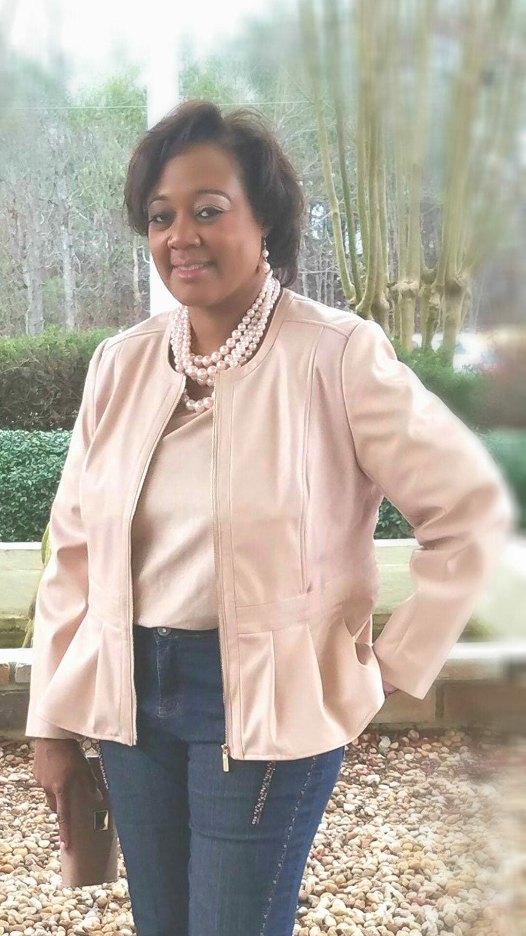 A Midnight Velvet customer in a blush top and peplum jacket set, jeans, a pearl necklace, and holding a blush clutch.