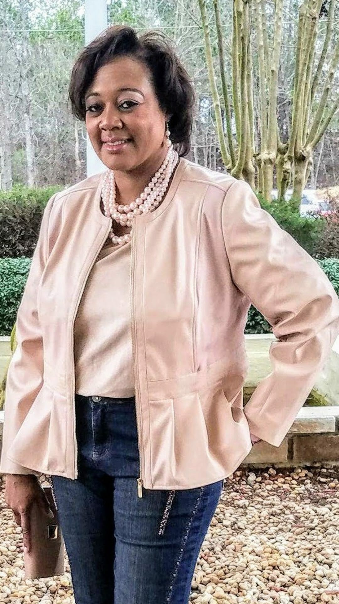 A Midnight Velvet customer in a blush top and peplum jacket set, jeans, a pearl necklace, and holding a blush clutch.