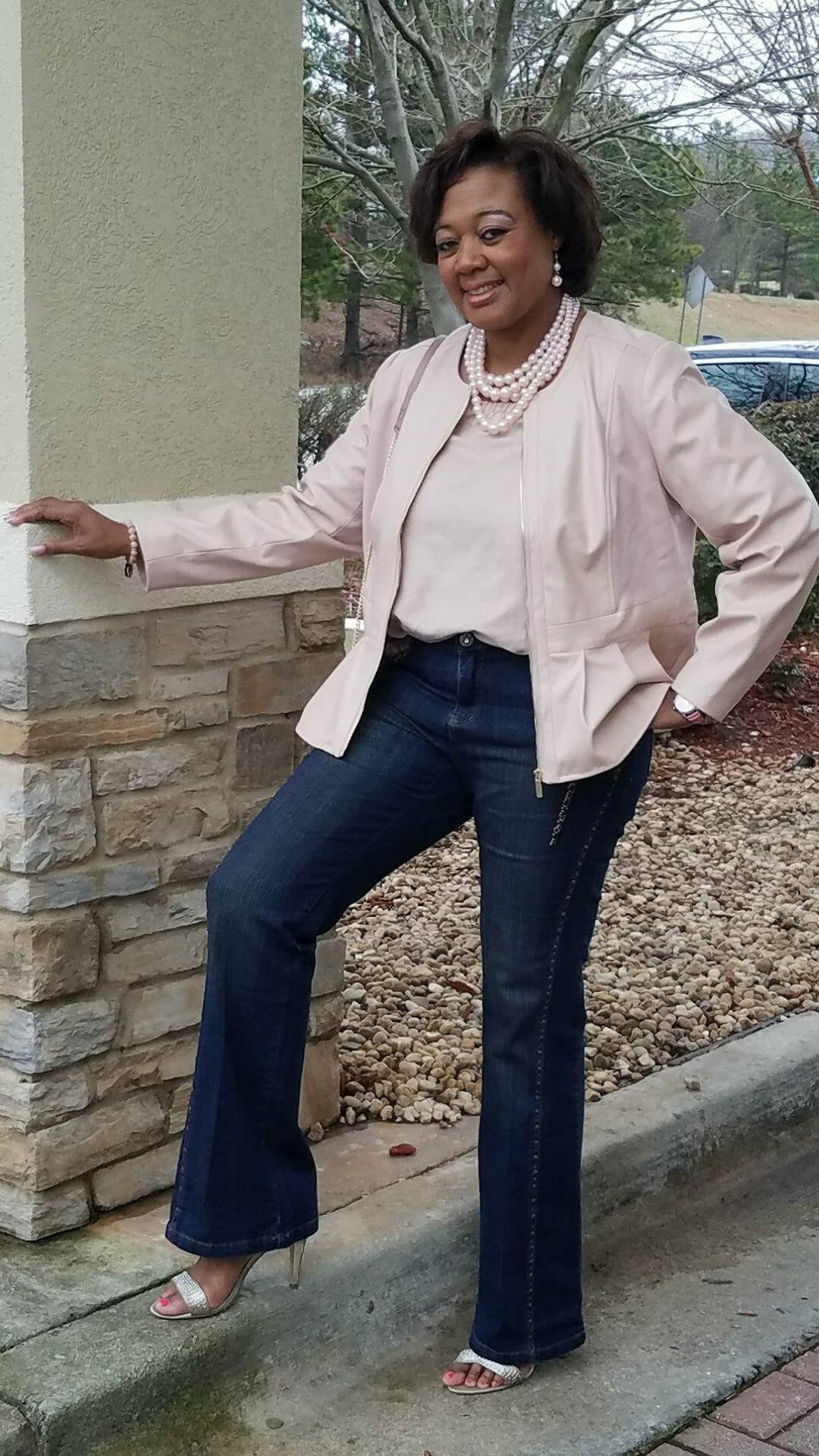 A Midnight Velvet customer by a rock column, in a blush top and peplum jacket set, jeans, sandals, and a pearl necklace.