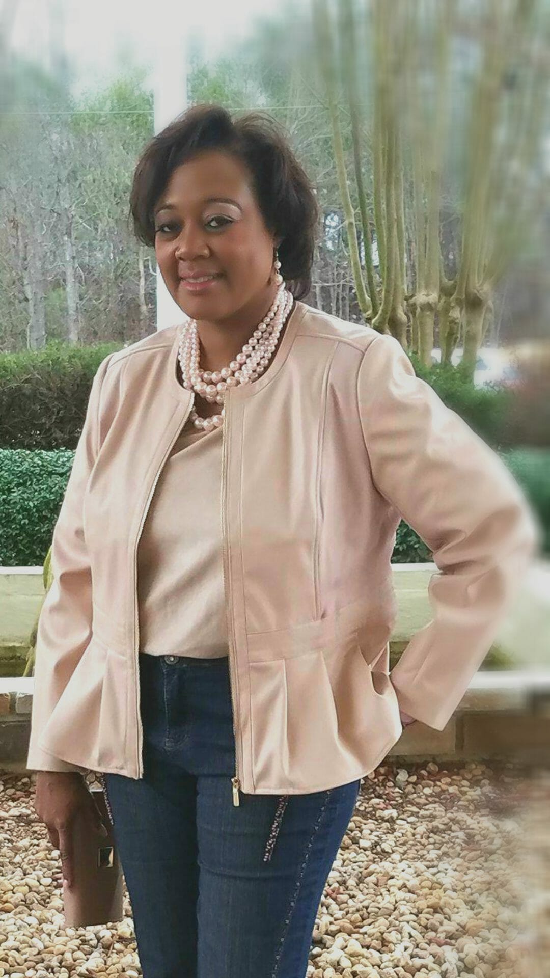 A Midnight Velvet customer in a blush top and peplum jacket set, jeans, a pearl necklace, and holding a blush clutch.