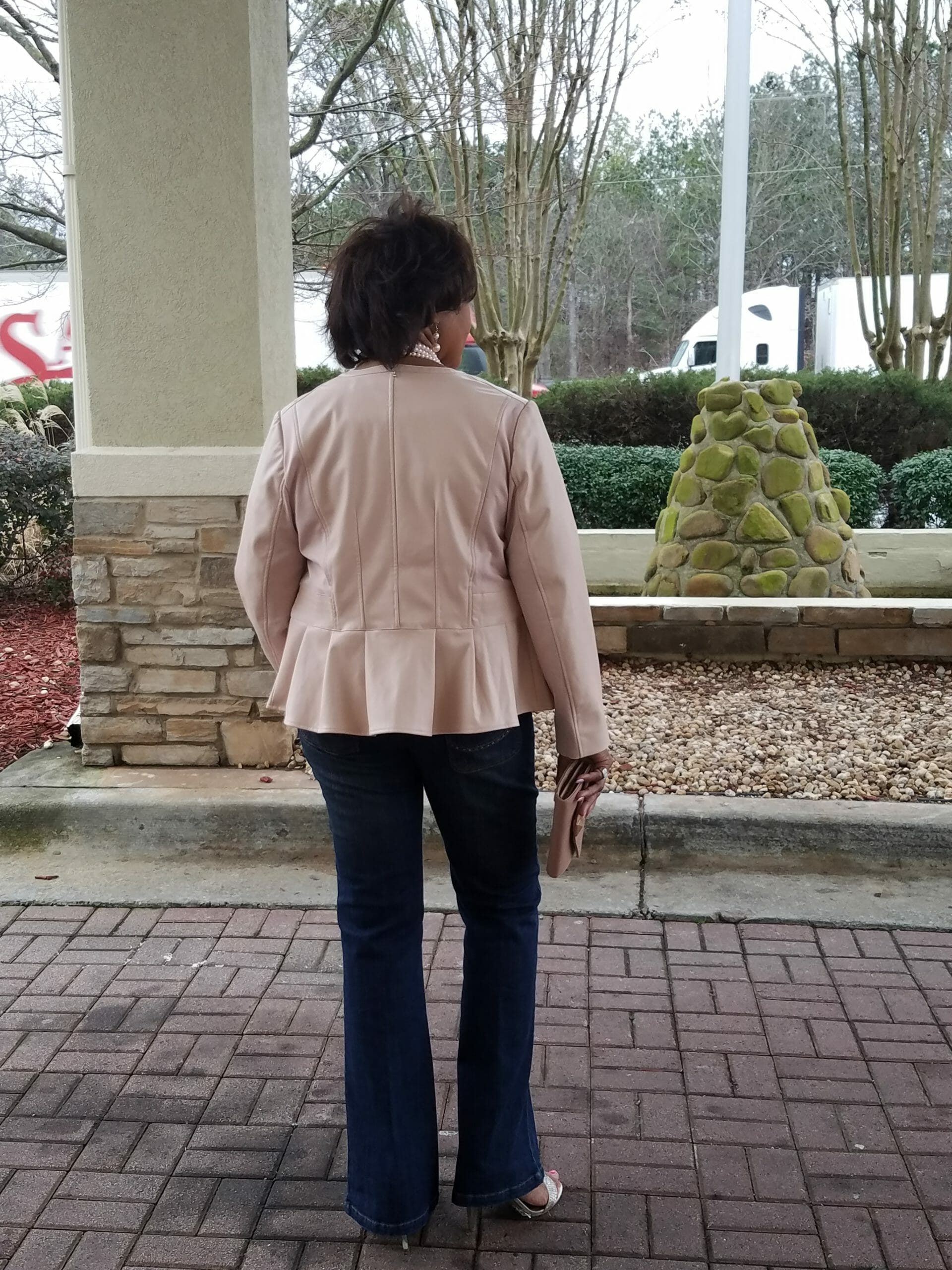 Back view of a Midnight Velvet customer outside in a blush peplum jacket, jeans, sandals, and a pearl necklace.