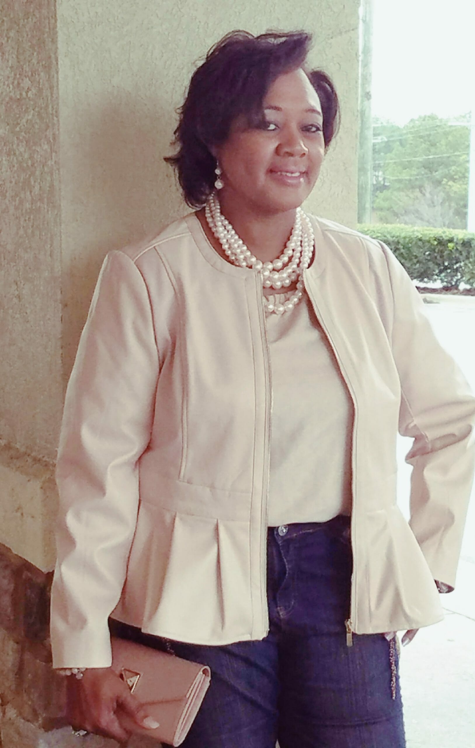 A smiling Midnight Velvet customer in a blush top and peplum jacket set, jeans, a pearl necklace, holding a blush clutch.