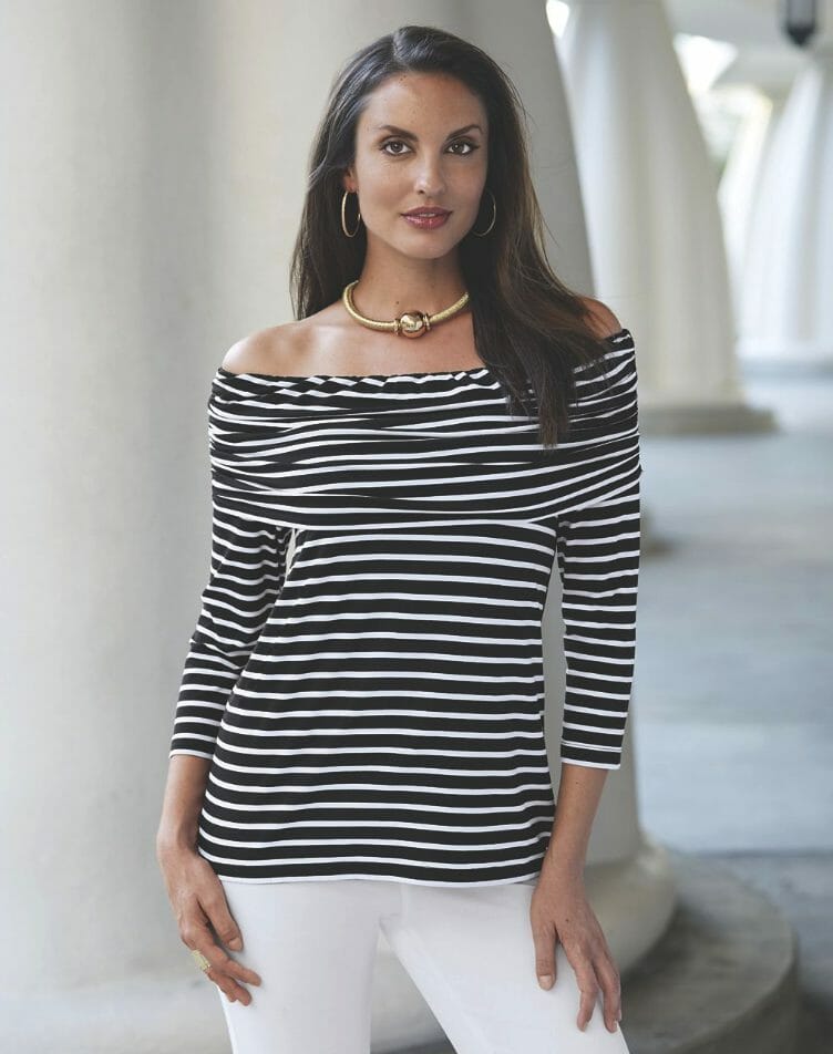 A brunette woman in a black and white stripe off-the-shoulder top, white pant, gold choker necklace and gold hoop earrings.