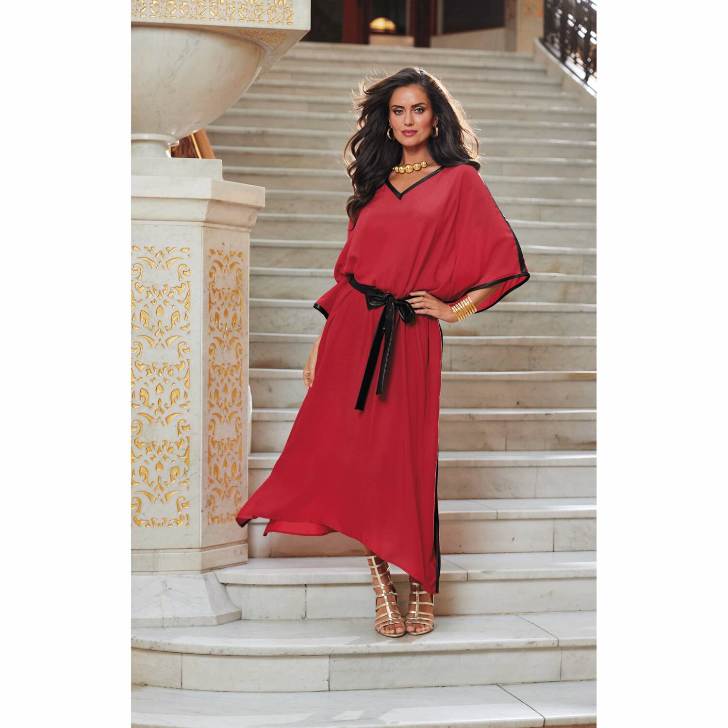 A woman in a flowing red dress with black accents stands confidently on a grand staircase, showcasing elegant accessories.