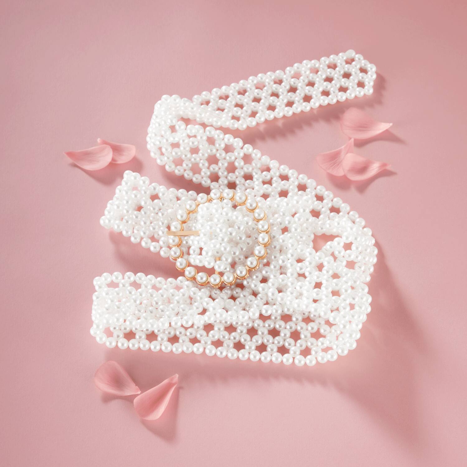 A delicate pearl belt with a circular pearl brooch, surrounded by soft pink petals on a pastel pink background.