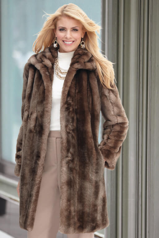 Smiling blonde woman wearing a long brown faux-fur coat with collar, white top, tan pant, and a gold chain necklace.