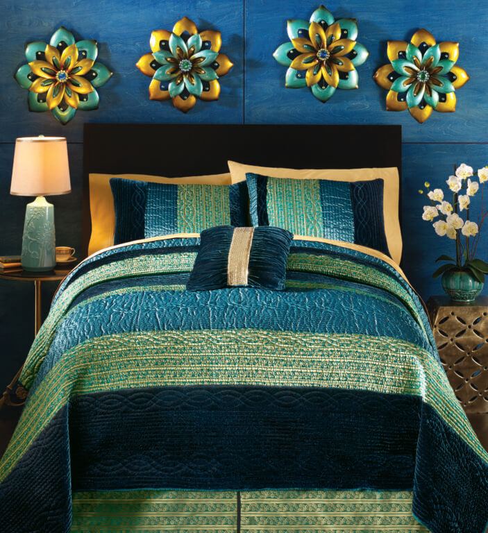 A velvet quilted bed set in shades of blue against a deep blue wall, with wall decor of four metallic blue and gold flowers.