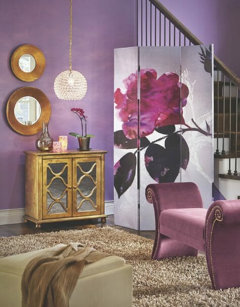 An area in shades of mauve, a bench, 3-panel screen of a large rose, gold glass door console and two round gold wall mirrors.