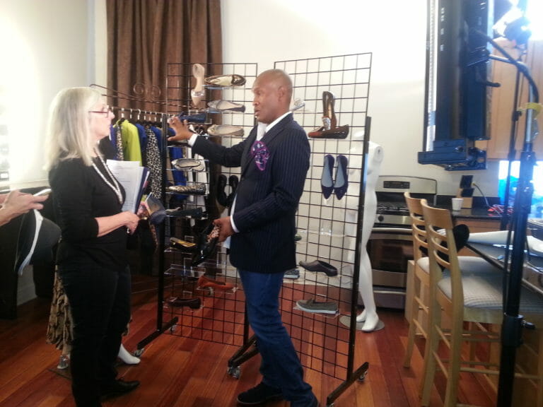 A designer black man in a suit coat, speaking to a white woman about a clothes and shoes video being shot in a kitchen.