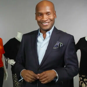 Marcellas Reynolds, a smiling designer black man in a suit jacket and white shirt, in front of dressed mannequins.