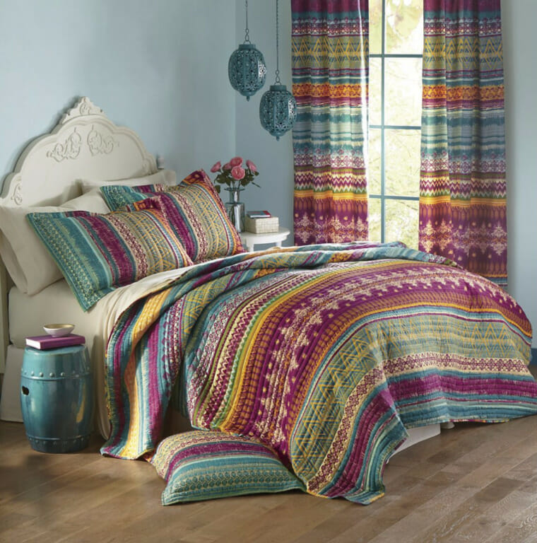 A Moroccan print bedding set in teals, gold and burgundy, with matching curtains and pillow, and a teal ceramic side table.