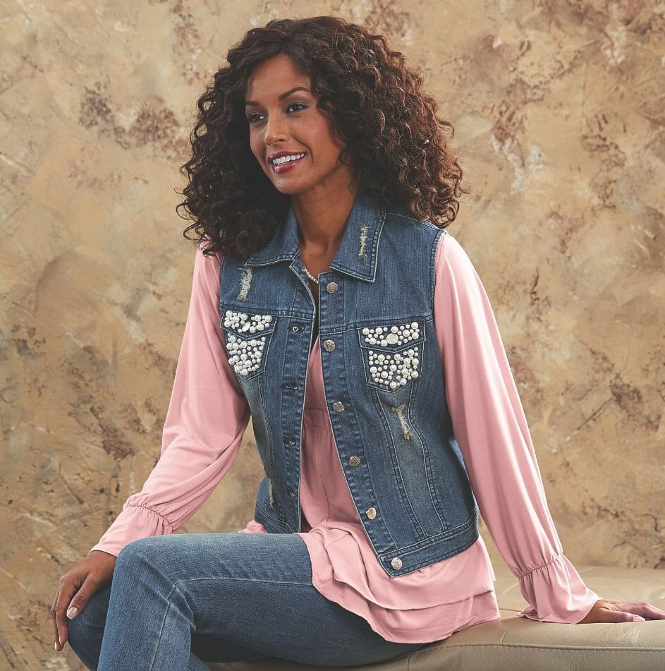 A smiling black woman wearing a denim vest with white embroidered chest pockets, a pink long-sleeved tunic, and jeans.
