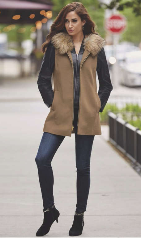 A woman on a sidewalk in a tan sleeveless vest with a faux fur hood over a black jacket, skinny jeans and black booties.