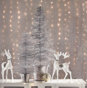 Two silver bottle brush trees by two white reindeer figurines, and a backdrop of sheer fabric over lit strings of lights.