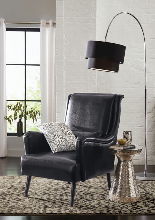 A black faux leather armchair with an animal print pillow, a chrome floor lamp with a black shade, and a copper side table.