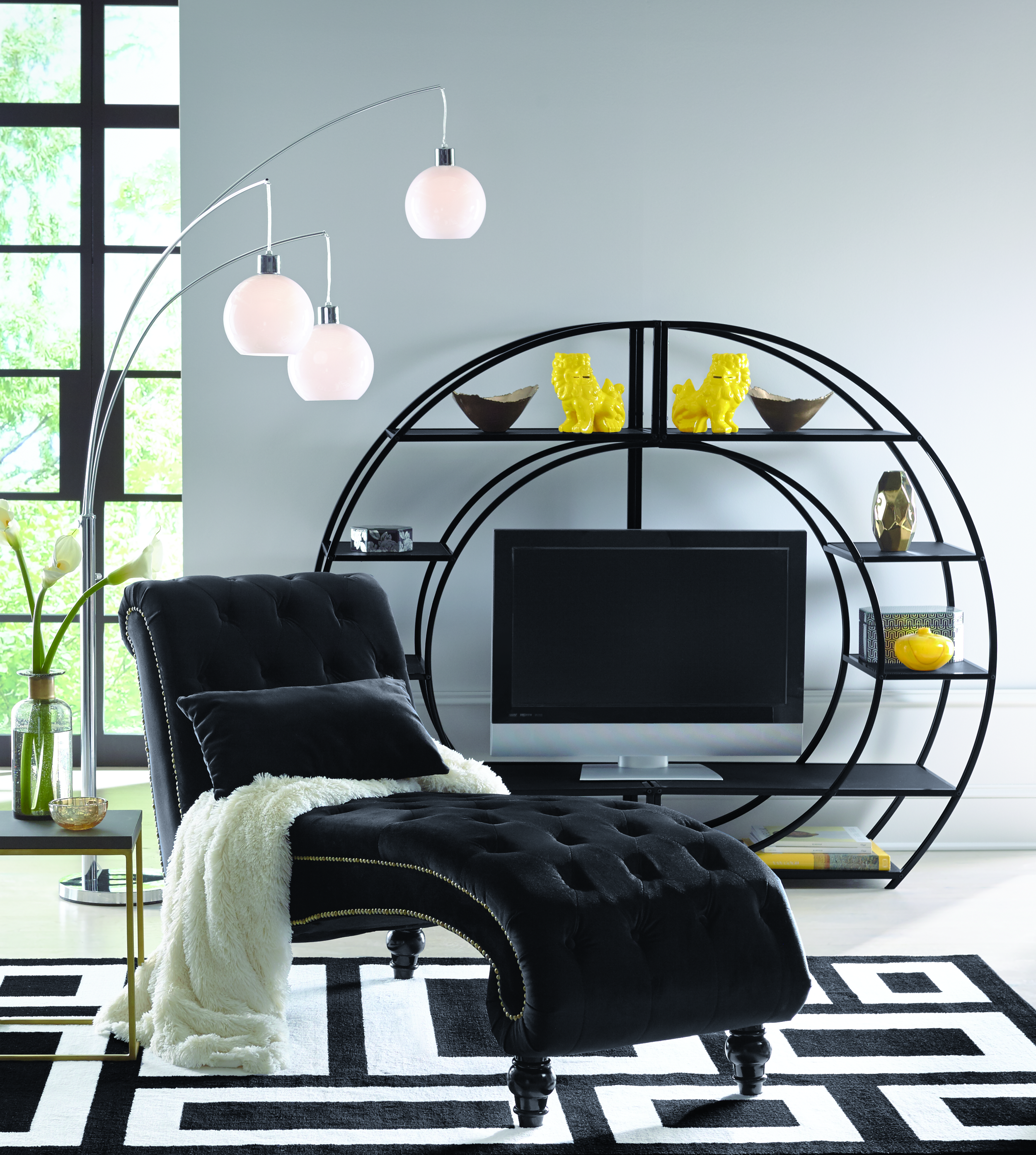 Black tufted chaise with a white plush throw, chrome floor lamp with three white globes, a black circle TV stand and shelves.