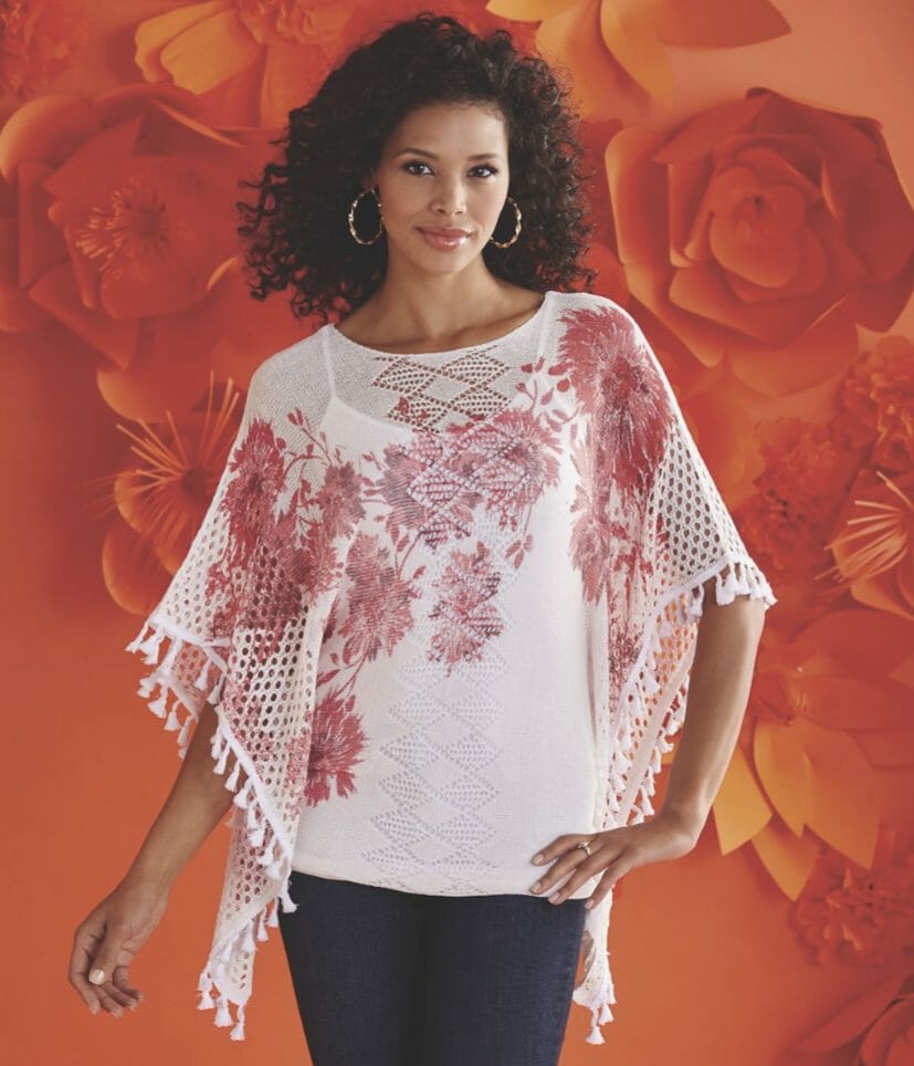 A black woman wearing a white cape-like top with crocheted and tasseled sleeves and a burgundy floral design.