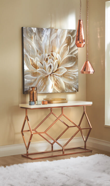 A large wall canvas of an ivory dahlia, a copper vase on a white and copper console, and two hanging copper lamps.