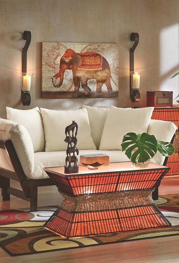 An Afrocentric living room with an ivory futon, lit orange and wicker tables, an elephant wall canvas and figurines.