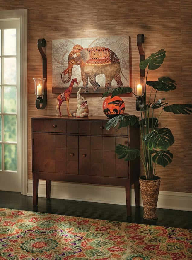 An orange and brown elephant canvas and two lit sconces above a wooden console with African figurines, and a green plant.