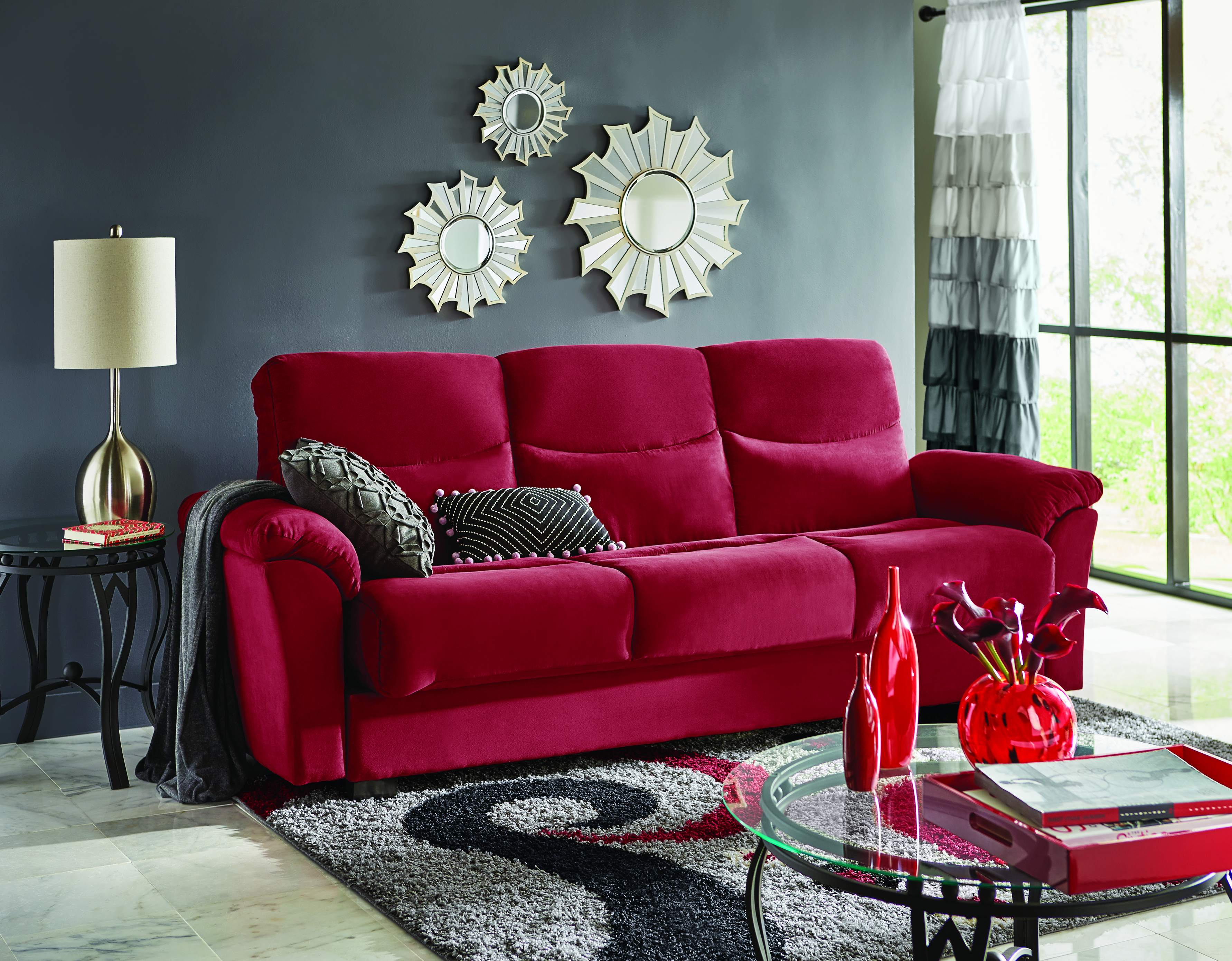 Red sofa with gray pillows and throw, three sunburst wall mirrors, a gray, black and red swirl rug, and glass topped tables.